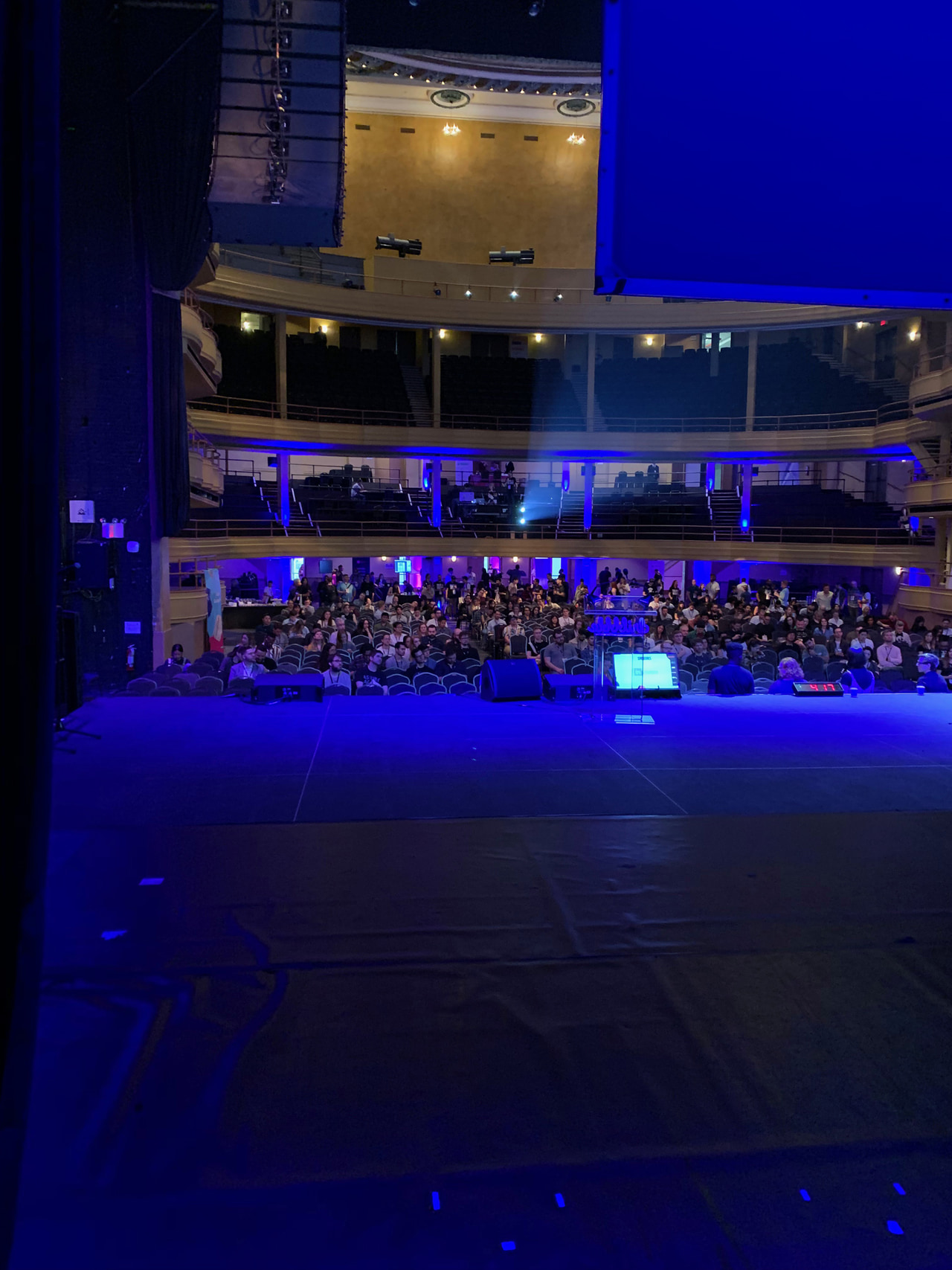 Blue-lit theater viewed from the stage, with an audience beneath curved balconies.