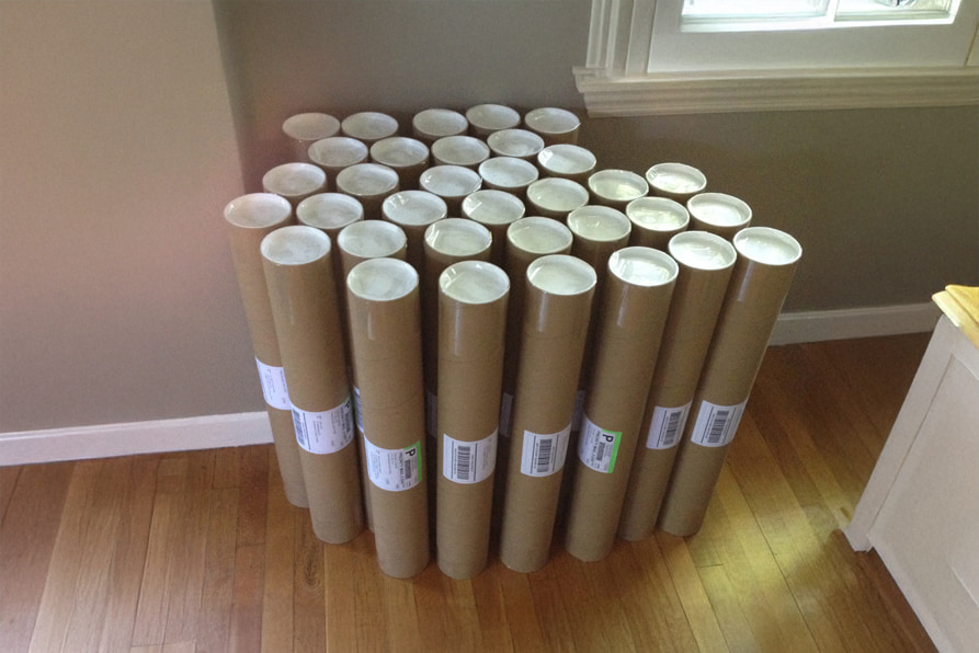 A group of tall cardboard mailing tubes with white caps and shipping labels standing on a wooden floor.