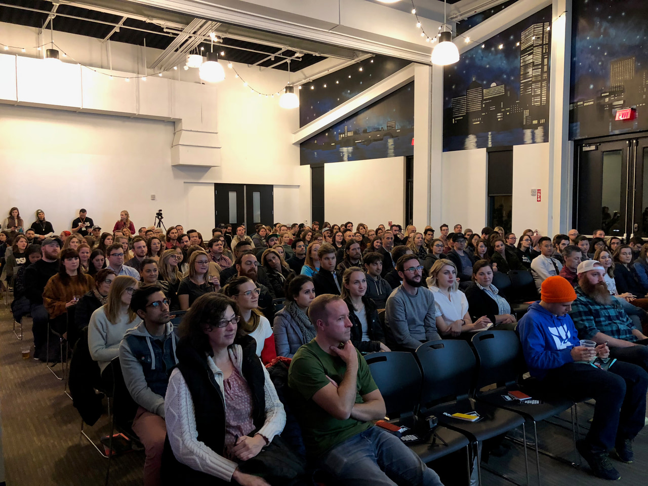 Large seated audience in a bright room with tall windows and exposed ceiling beams.