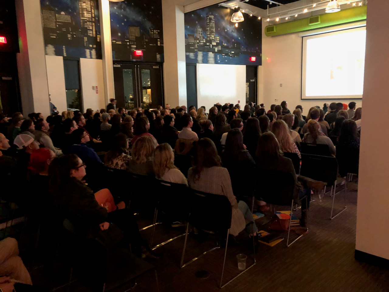 Audience facing a projected presentation in a room with tall windows and exposed ceiling beams.