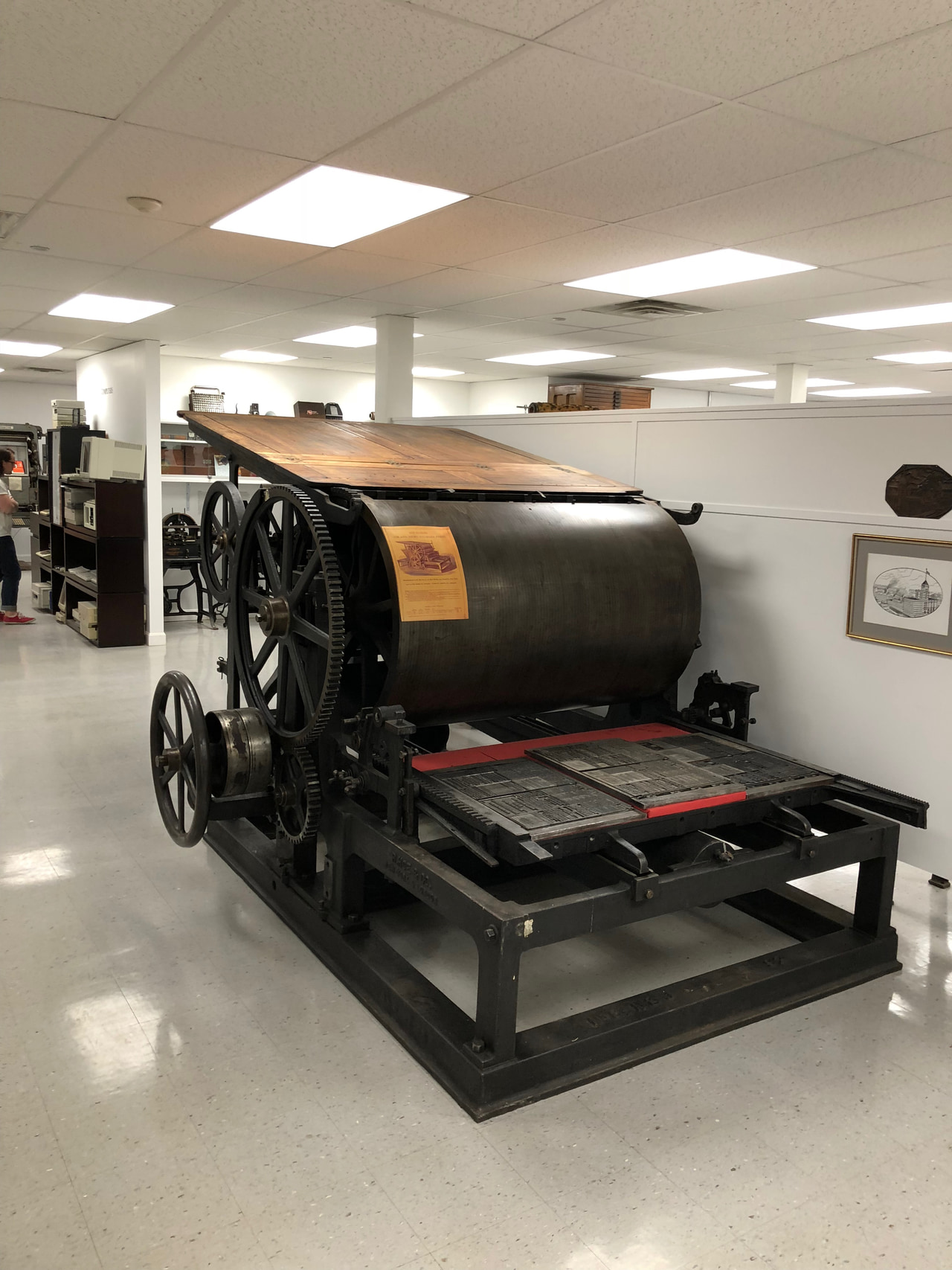 Large vintage printing press with a black roller and flywheel in a brightly lit museum room.