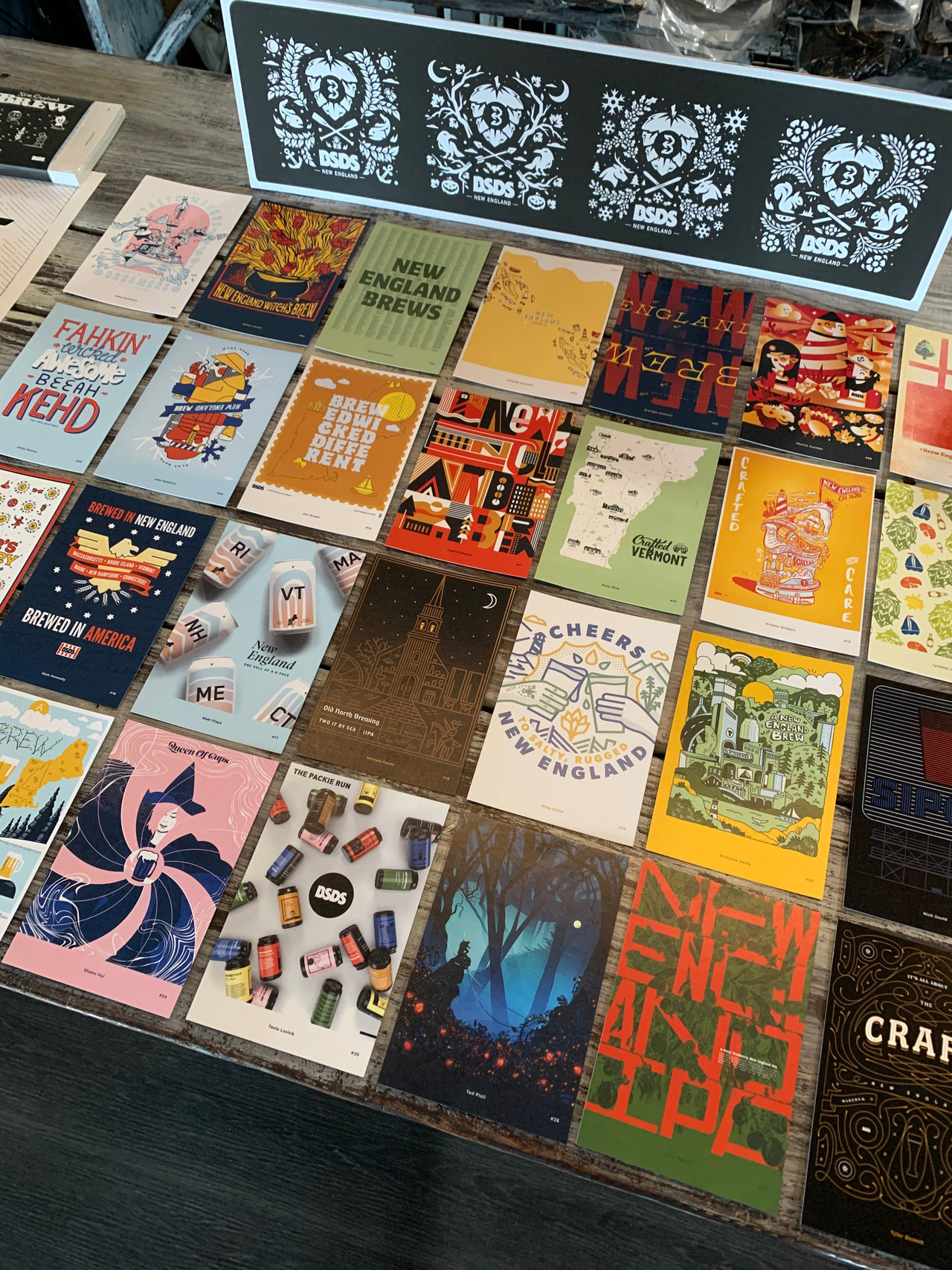 Table display of colorful posters, prints and publications, with a black-and-white ornamental print propped at the back.