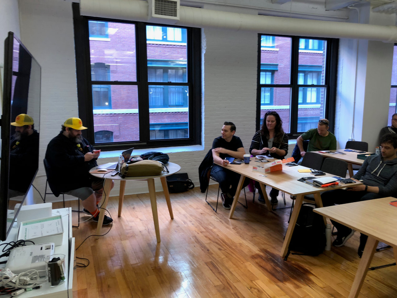 Small group reviewing work around a table beside large windows, with a presenter in a yellow cap at the left.