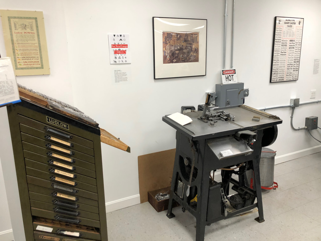 Printing press and wooden type cabinet displayed in a museum room beneath framed prints.