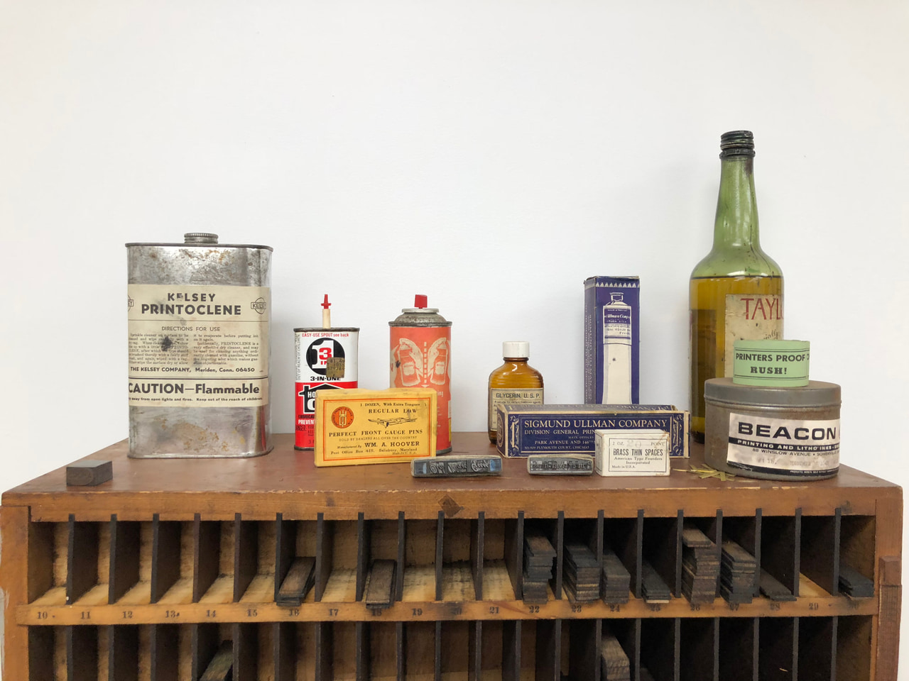Vintage printing supplies, including metal solvent and oil containers and a green bottle, displayed above a wooden type cabinet.