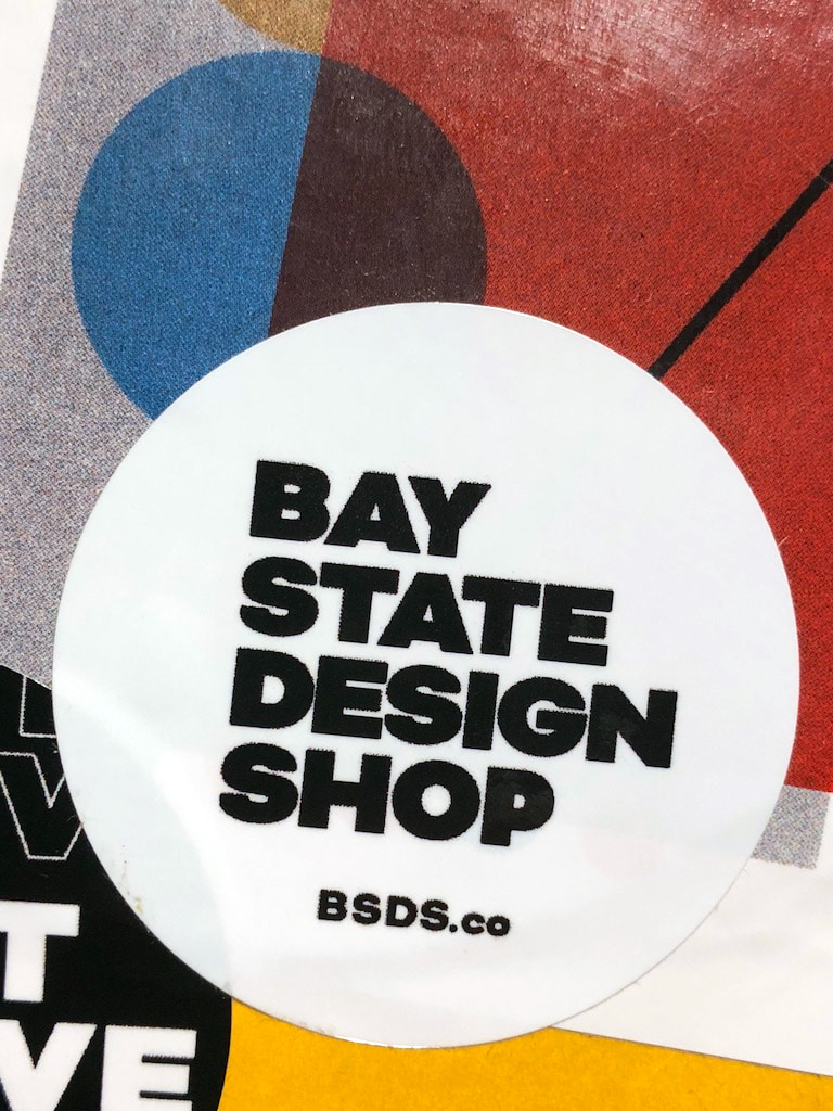 Round white Bay State Design Shop sticker with bold black lettering and BSDS.co, over a colorful printed background.