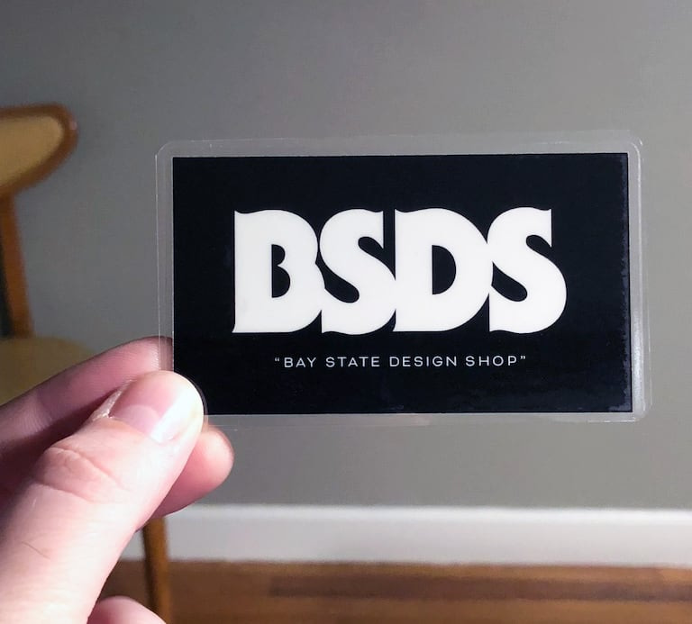 Hand holding a black BSDS sticker with large white lettering and Bay State Design Shop written underneath.