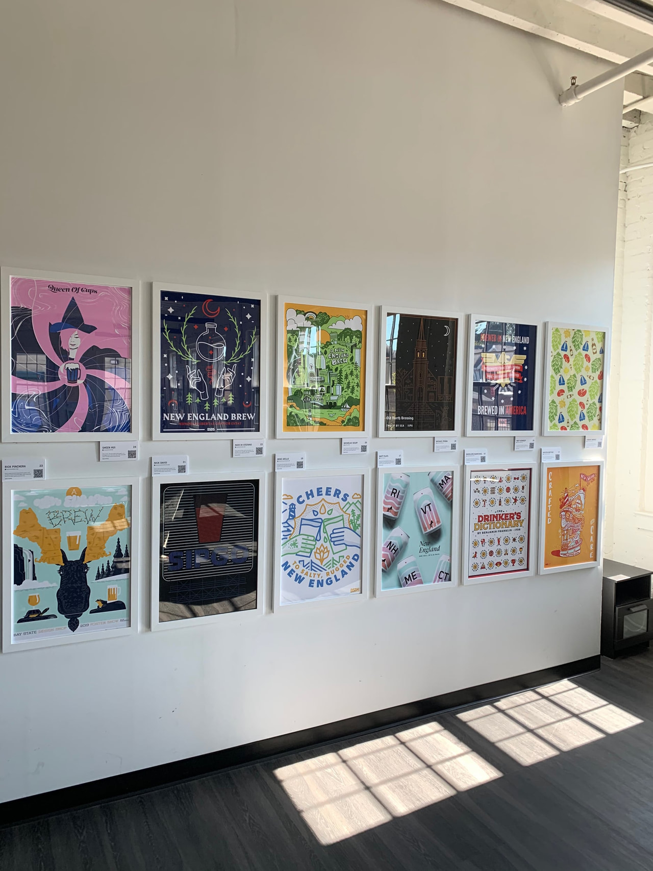 Colorful posters displayed in two rows along a white gallery wall, with sunlight across the floor.
