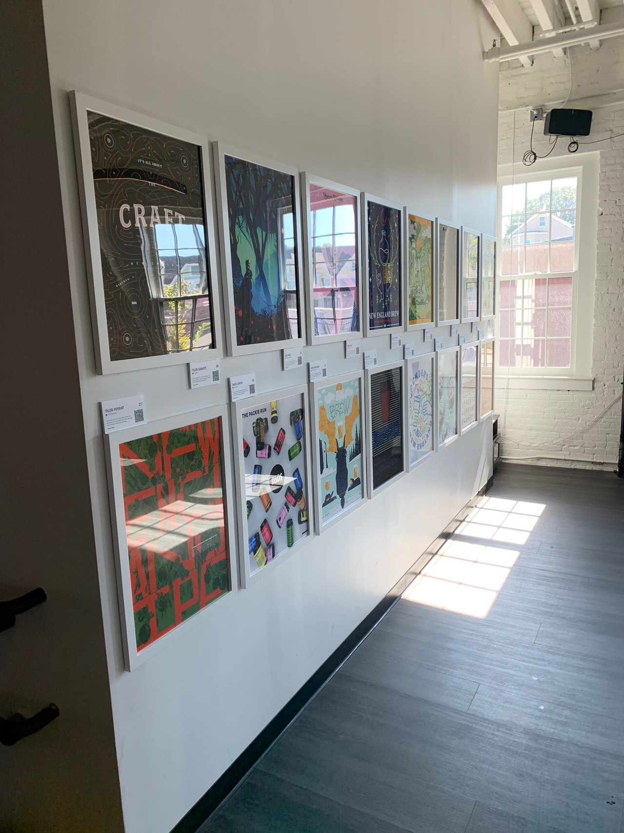 Two rows of posters along a white gallery wall beside tall windows and a sunlit floor.