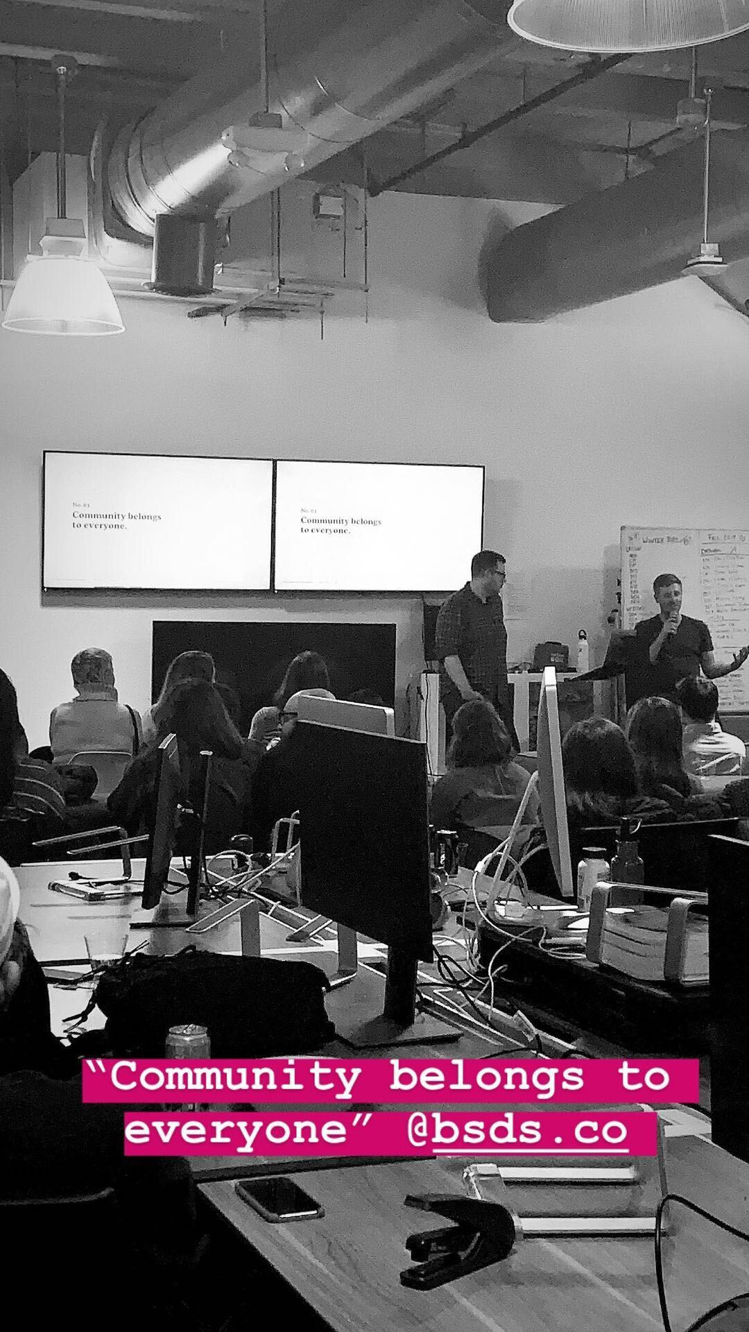 Black-and-white photograph of a workshop audience with pink caption text: Community belongs to everyone, @bsds.co.