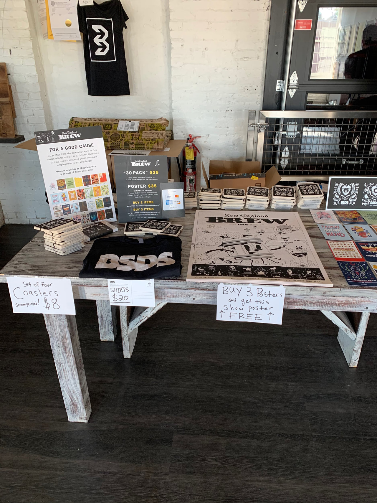 A rustic wooden table displays merchandise including t-shirts, coasters, posters, and a large illustrated poster. A white brick wall and a metal door are in the background. Handwritten signs indicate prices and promotions.