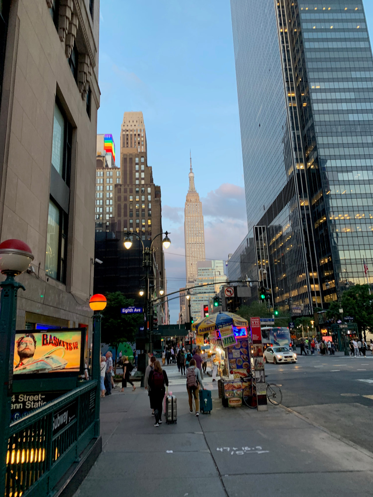 City street lined with tall buildings, pedestrians, sidewalk vendors and a subway entrance.