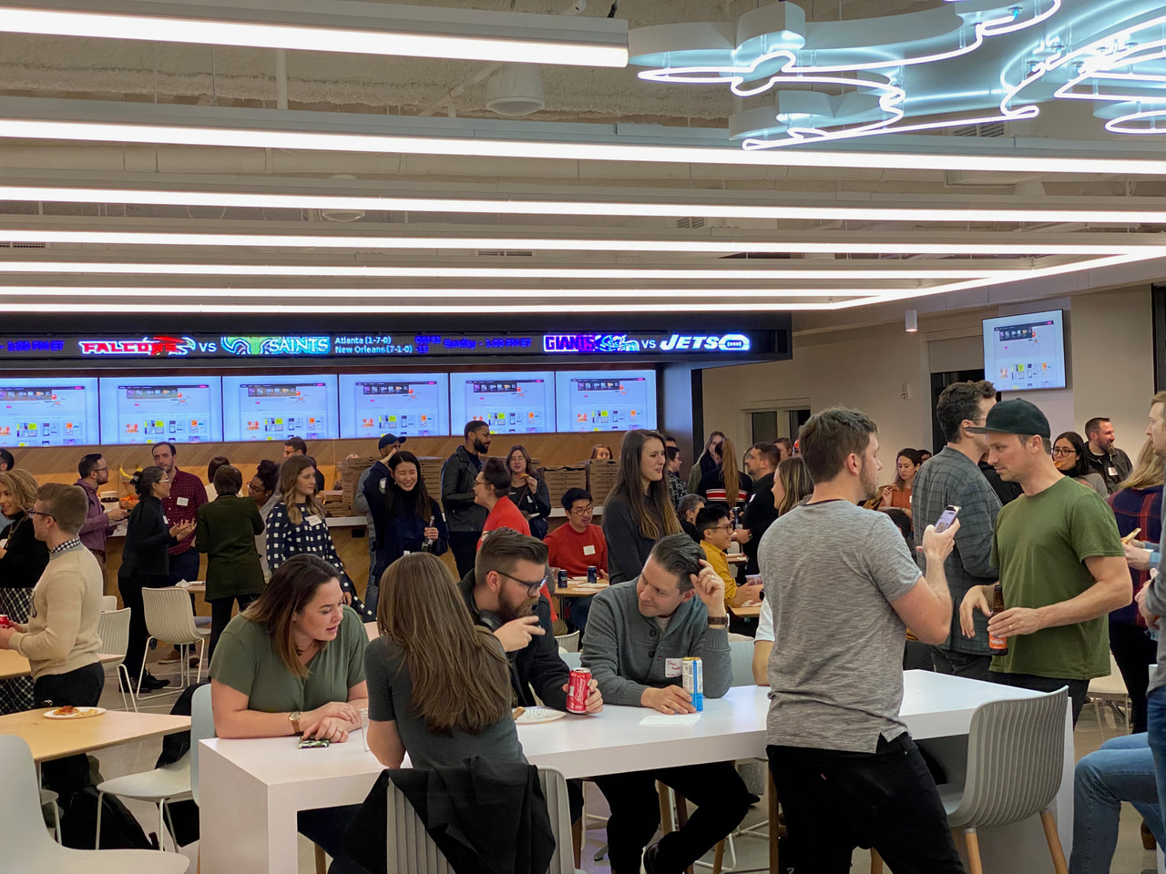 People talking around long tables beneath linear ceiling lights and blue neon lettering.