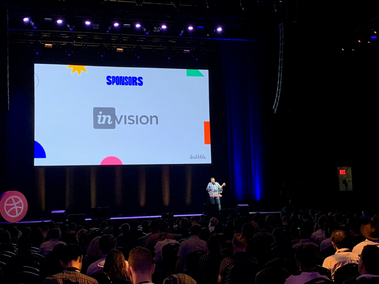 A speaker stands below a projected sponsor slide in a dark auditorium.