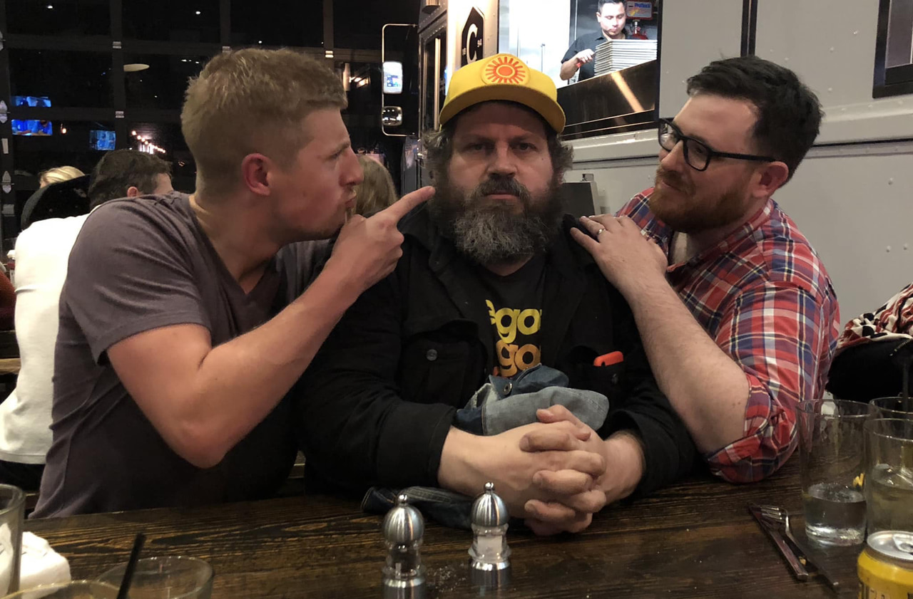 Three people pose together at a table, with a bearded person seated in the center.