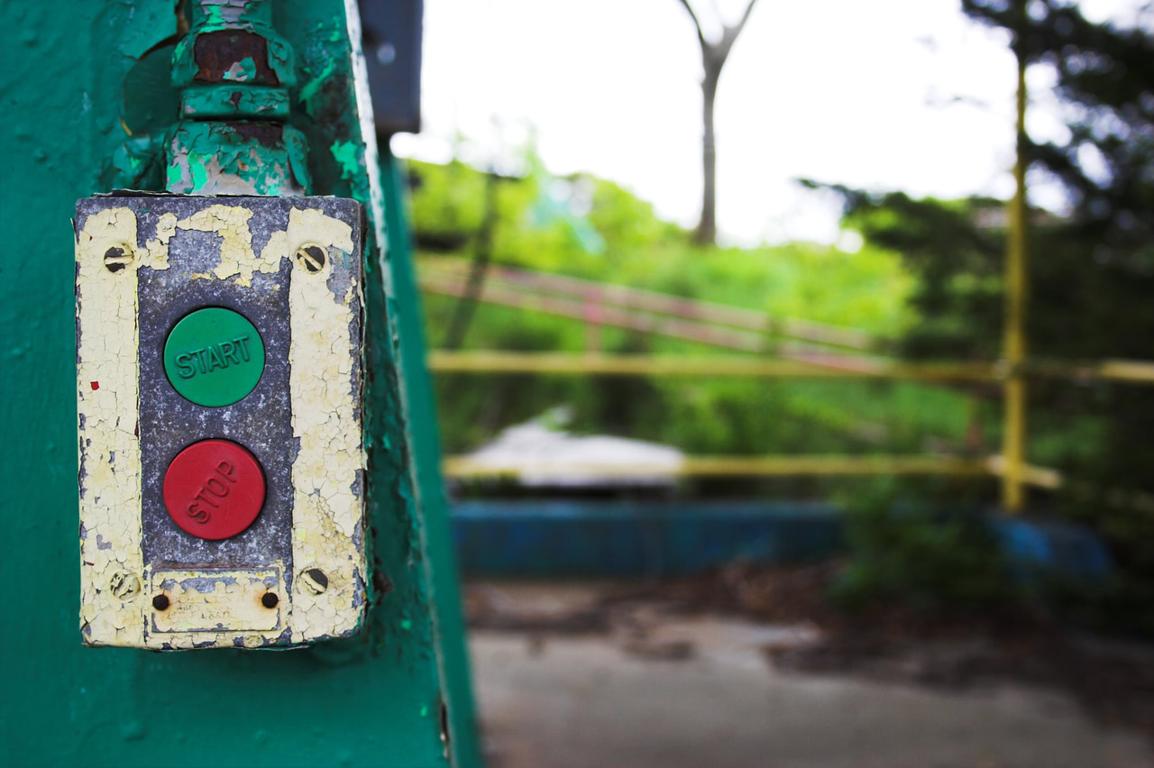Weathered green Start and red Stop buttons on a peeling metal post, with a blurred fence and trees behind.