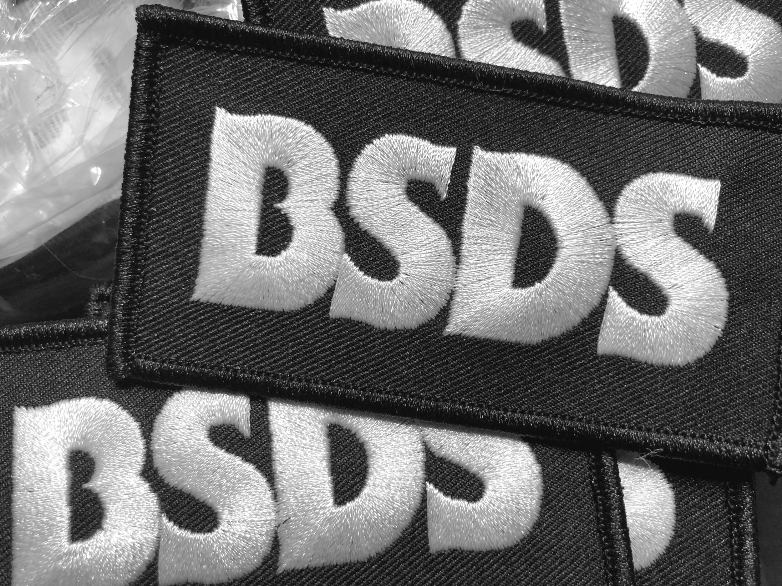 Close-up black-and-white photograph of embroidered BSDS patches with raised white lettering on black.