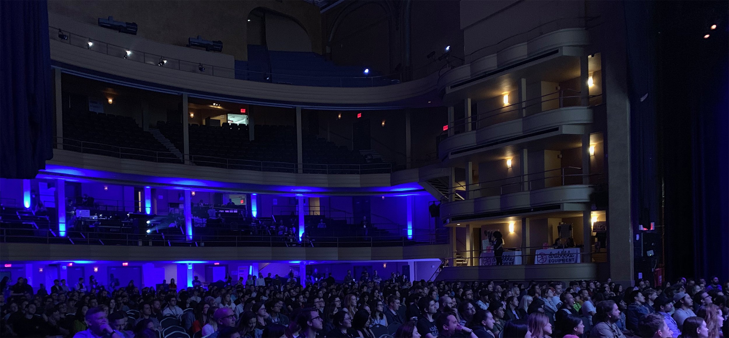 Audience in a theater with curved balconies and blue lighting.